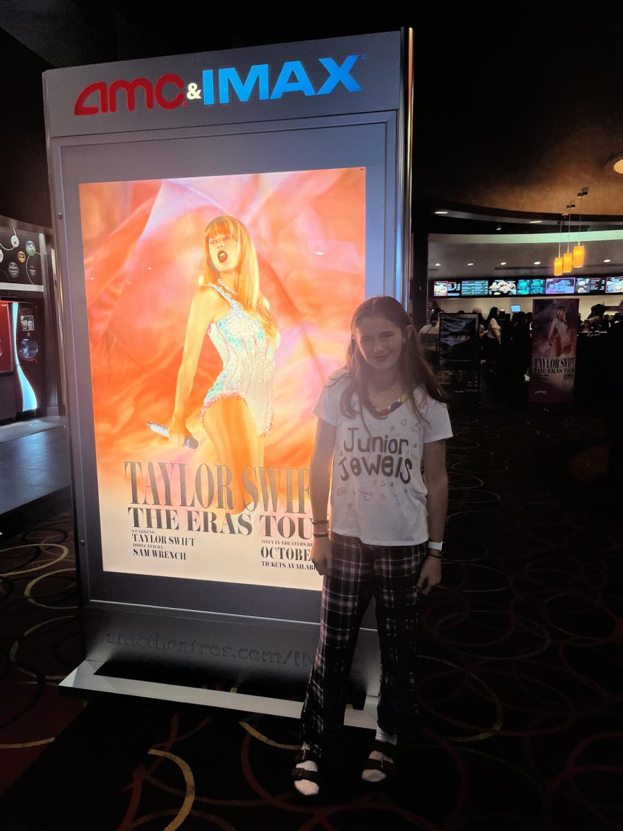 Staff Writer Clio Long poses next to the Eras Tour sign on opening day. She is wearing a shirt similar to the outfit that Taylor wears in her “You Belong With Me” music video.